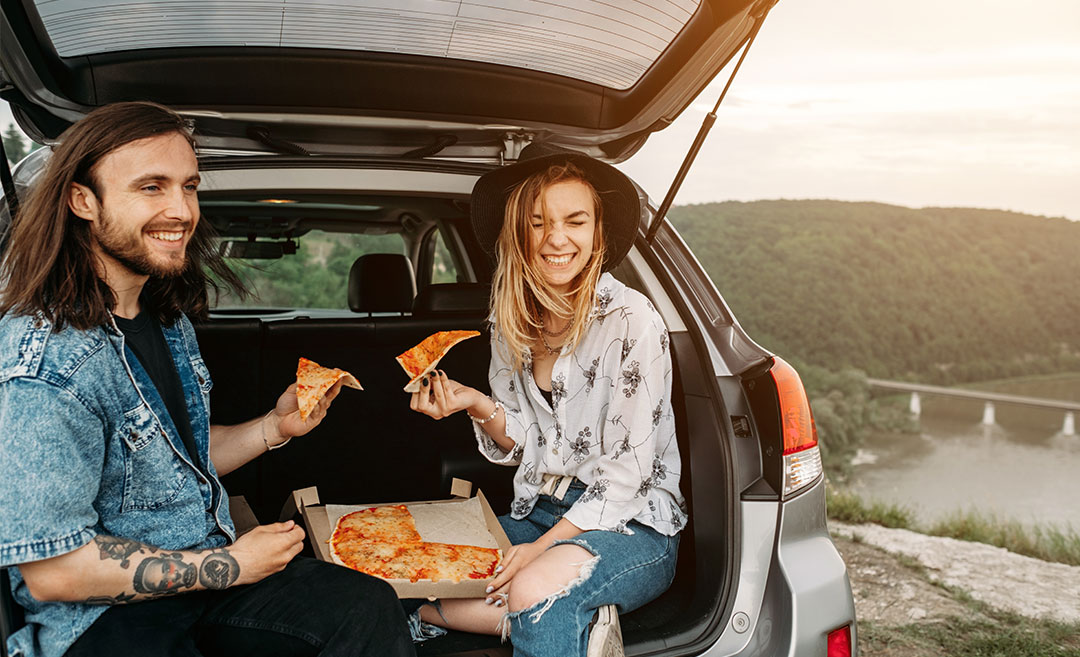 A young man and woman having fun sitting in a car trunk and eating pizza, travelling and road trip.