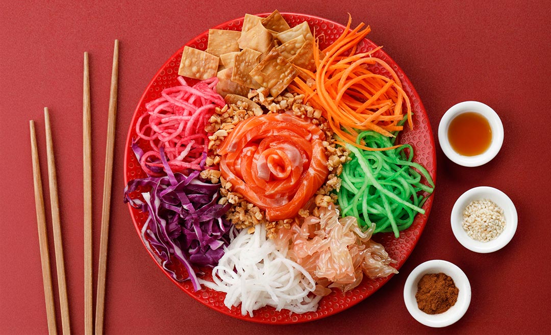 An editorial flat lay of a colourful Yee Sang prosperity toss plate with raw salmon, shredded vegetables, chopsticks, and small bowls of plum sauce and sesame oil.