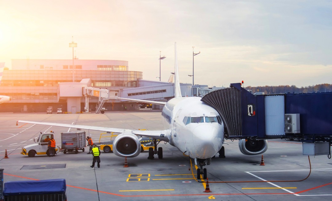 An airplane passenger gangway connected to an aircraft at a Malaysian airport terminal, with flight maintenance crews preparing for a domestic departure.