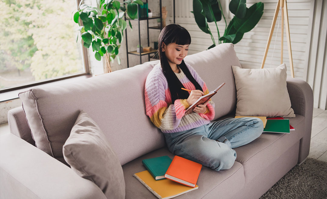 A young woman sitting comfortably on a sofa in a bright, cosy living room, smiling as she writes in her travel journal.