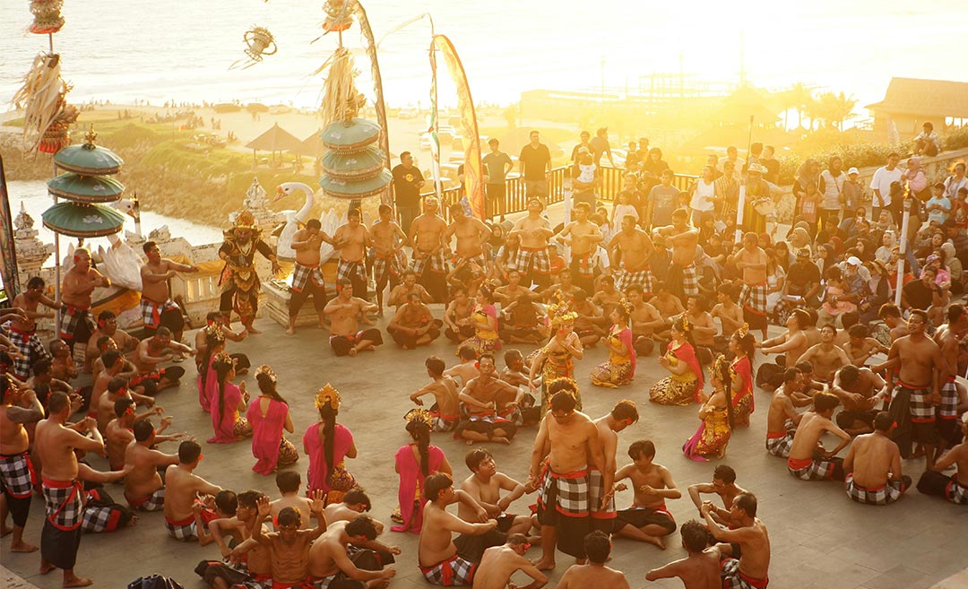 A traditional Balinese Kecak Dance performance at sunset in Bali, Indonesia, featuring performers in a circle against a dramatic twilight sky.