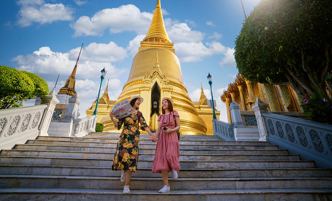 Two female friends walking together up the decorative stone steps of a traditional Thai temple in Thailand, illustrating regional leisure travel and cultural sightseeing.