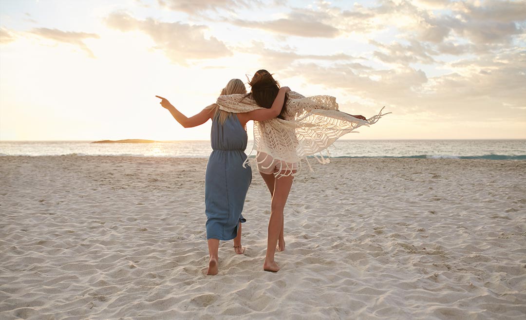 Two women walking along a sandy beach towards the sea with their arms around each other's shoulders during a trip.
