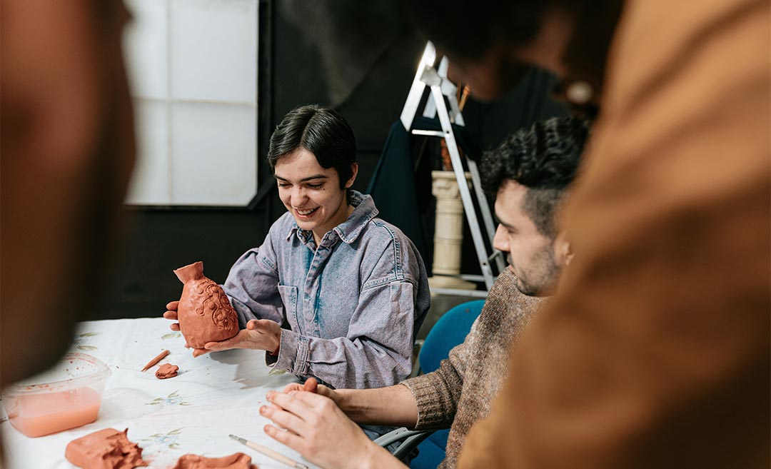 Diverse group of people sitting around a wooden table crafting pottery and shaping clay by hand in a workshop.