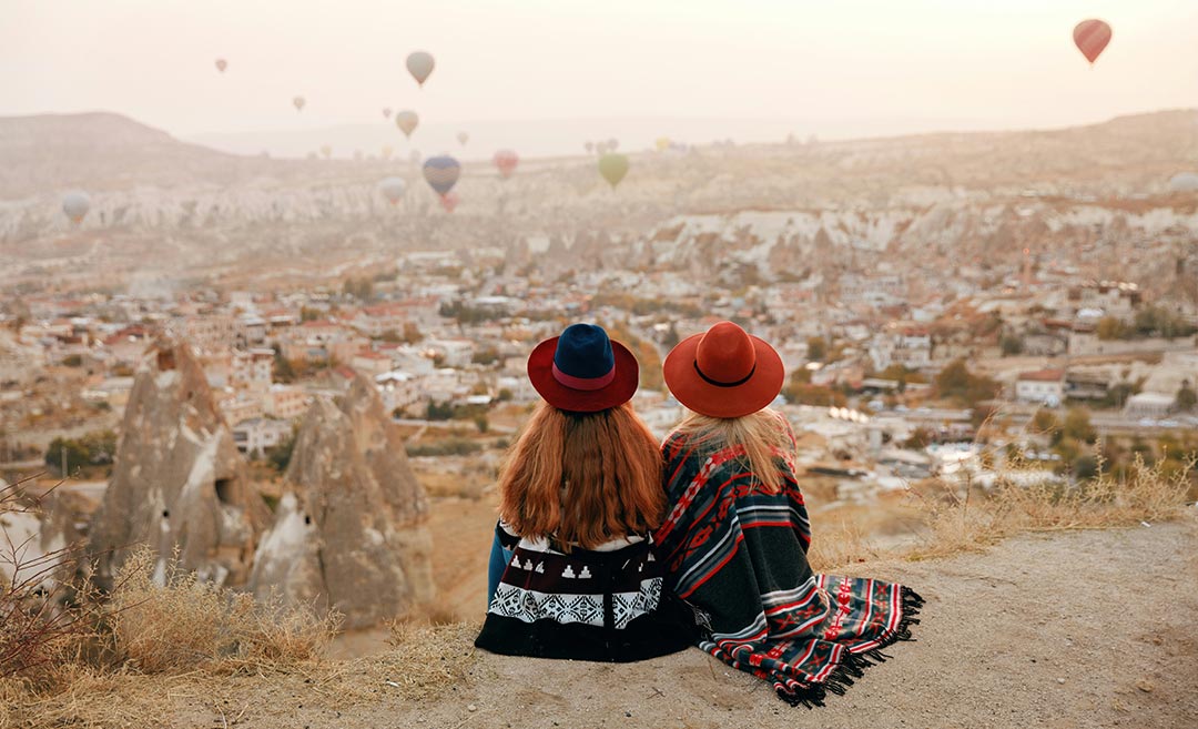 Two women wearing hats sitting on a hillside, enjoying the panoramic view of hot air balloons flying over Cappadocia, Turkey.