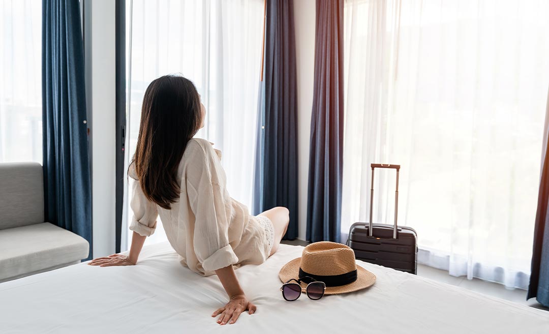 A relaxed woman traveller sitting comfortably on a hotel bed with her luggage unpacked beside her.