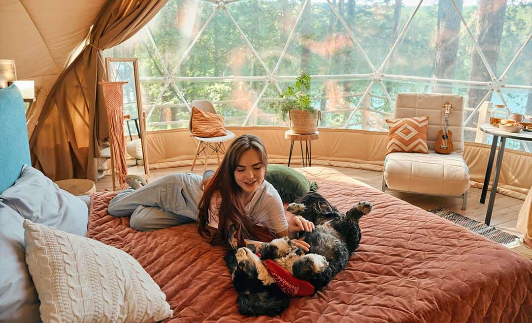 A woman and her dog relaxing comfortably inside a glowing geodesic igloo glass cabin at a glamping site in the woods.