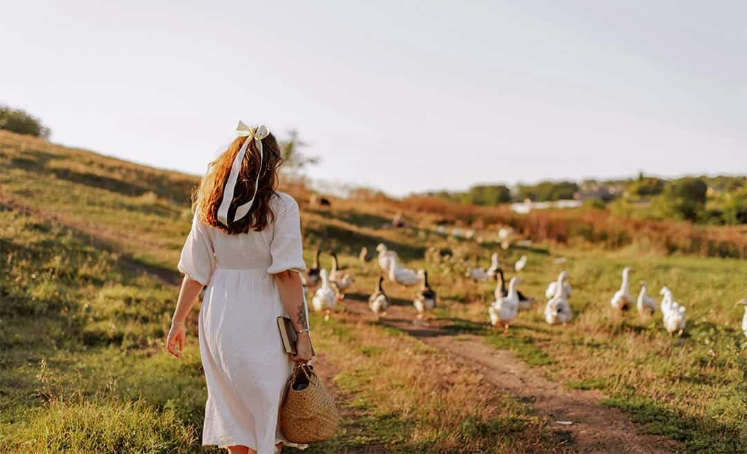 A woman in a white dress walking down a rustic country dirt road with geese foraging in the foreground.
