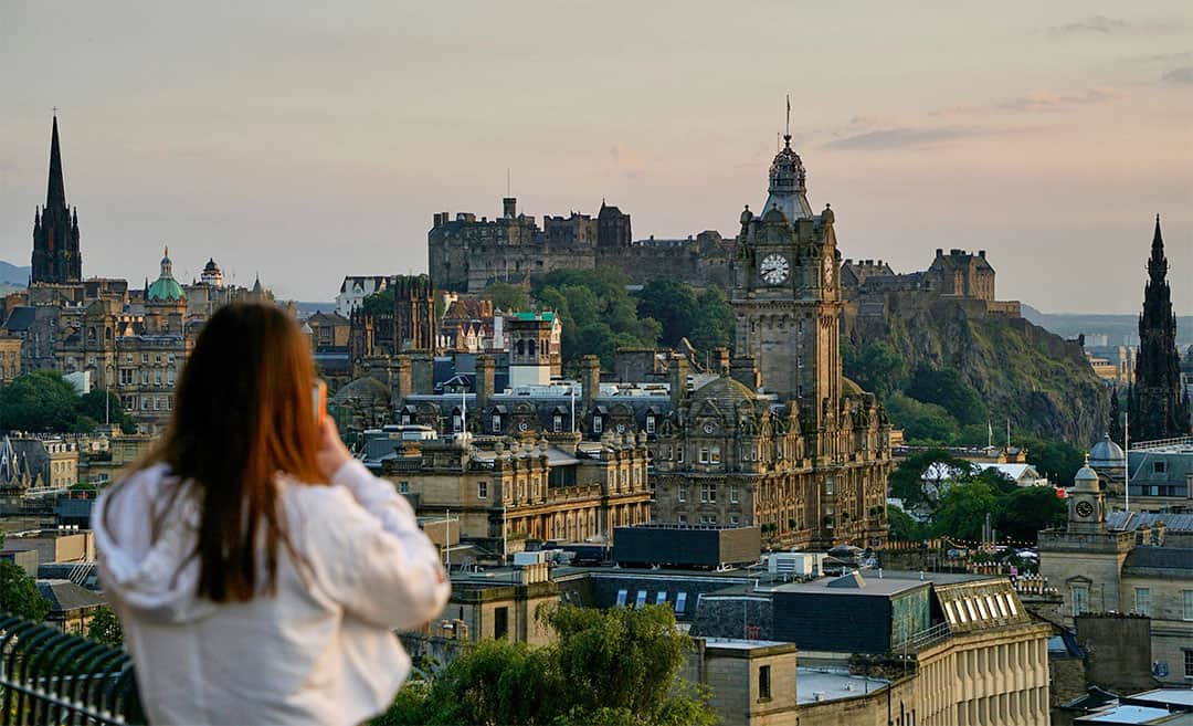 A woman in a white shirt looking out over the Edinburgh skyline from Calton Hill, with the Balmoral Hotel tower and Castle Hill visible in the background.
