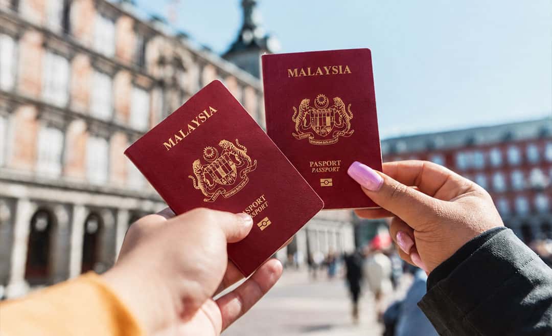 Two women holding up red Malaysian passports against a blurred outdoor background in Madrid, Spain.