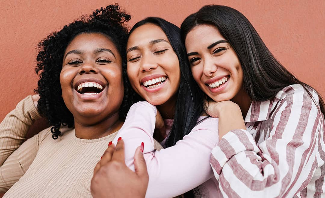 Three midlife female friends standing close together, smiling and embracing warmly on a trip.